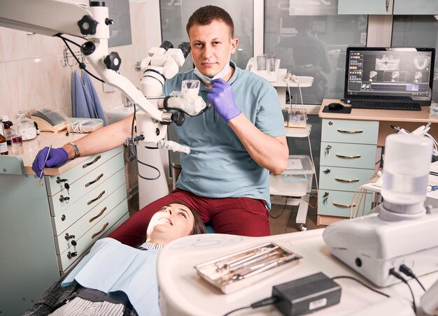 Joven dentista masculino sentado junto a la cama del paciente en el consultorio dental