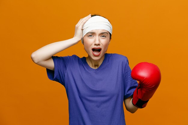 Joven deportista adolorida con camiseta y guante de boxeo mirando a la cámara manteniendo la mano en la cabeza con la cabeza envuelta con una venda aislada en el fondo naranja