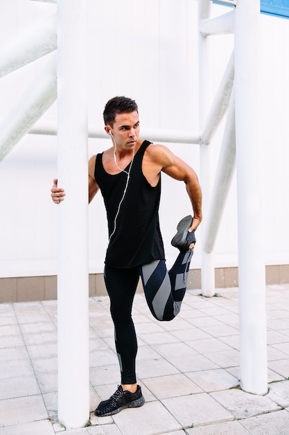 Joven deportista atractivo en auriculares haciendo ejercicios de estiramiento de deportes durante el entrenamiento