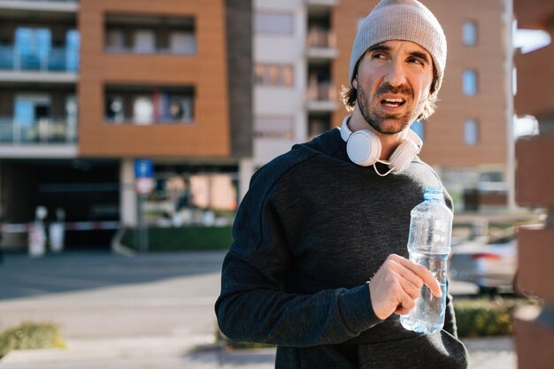 Joven deportista cansado con botella de agua tomando un descanso mientras hace ejercicio en la ciudad