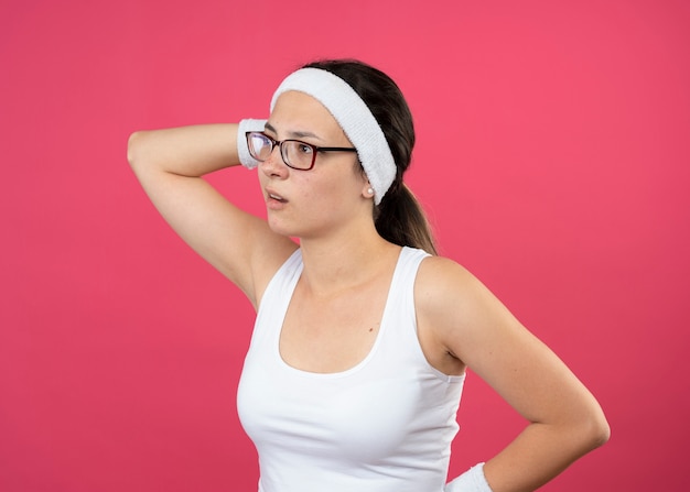 Joven deportista despistada con gafas ópticas con diadema y muñequeras sostiene la cabeza y mira el lado aislado en la pared rosa