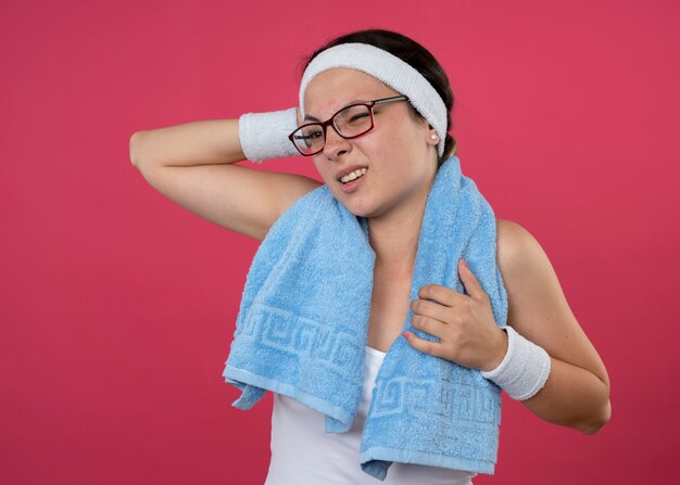 Joven deportista disgustada con gafas ópticas con una toalla alrededor del cuello con diadema y muñequeras pone la mano en la cabeza