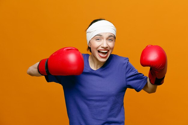 Joven deportista enojada con camiseta y guantes de boxeo mirando a la cámara manteniendo los puños en el aire con la cabeza envuelta con una venda aislada en el fondo naranja