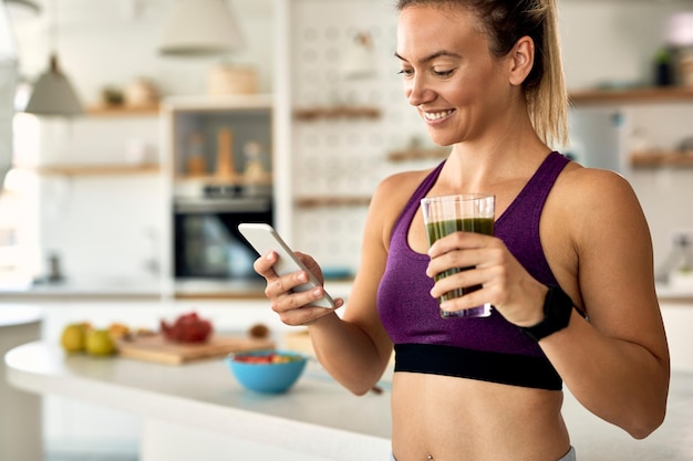 Joven deportista feliz usando un teléfono inteligente mientras bebe un batido de desintoxicación en la cocina