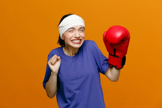 Joven deportista irritada con camiseta y guante de boxeo mirando al costado manteniendo los puños en el aire con la cabeza envuelta con una venda aislada en el fondo naranja