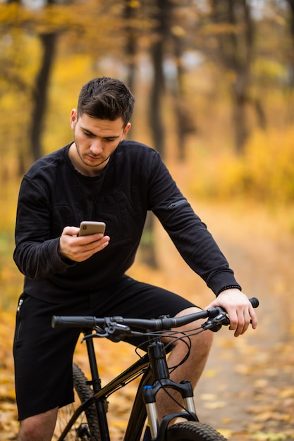 Joven deportista montando bicicleta sosteniendo el teléfono, soleado parque de otoño