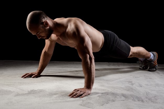 Joven deportista poderoso entrenamiento flexiones sobre la pared oscura.
