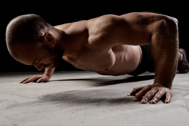 Joven deportista poderoso entrenamiento flexiones sobre la pared oscura.