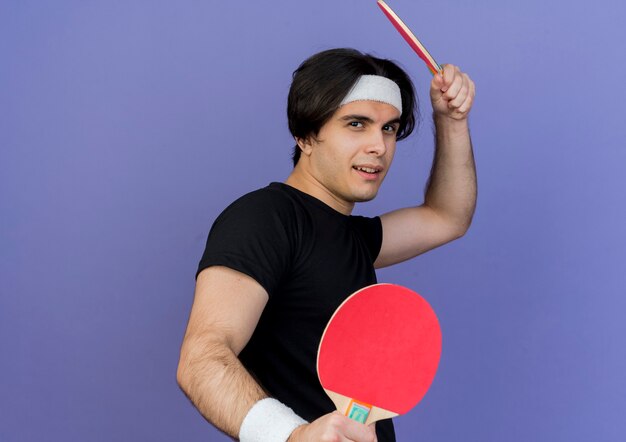 Joven deportista con ropa deportiva y diadema sosteniendo raquetas de tenis de mesa con cara seria