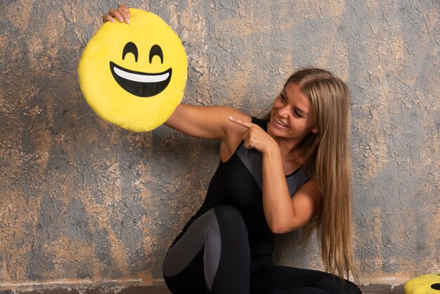 Joven deportista en ropa deportiva sosteniendo una almohada emoji sonriente y apuntando a ella.