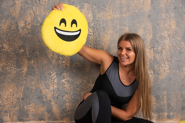 Joven deportista en ropa deportiva sosteniendo una almohada emoji sonriente arriba.