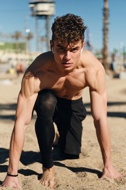 Joven deportista seguro de sí mismo mirando agresivamente a la cámara preparándose para correr al aire libre. Chico atractivo en entrenamiento en la playa de la ciudad