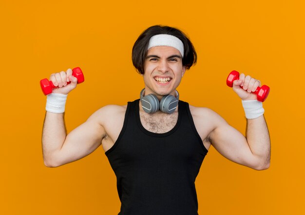 Joven deportista vistiendo ropa deportiva y diadema con auriculares alrededor del cuello trabajando con pesas sonriendo alegremente