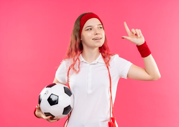 Joven deportiva en diadema sosteniendo un balón de fútbol apuntando hacia arriba con el dedo índice mirando confiado de pie sobre la pared rosa