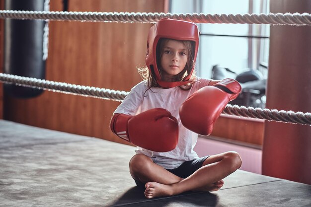 Una joven deportiva está sentada en un anillo soleado usando su equipo: casco y guantes de boxeo.