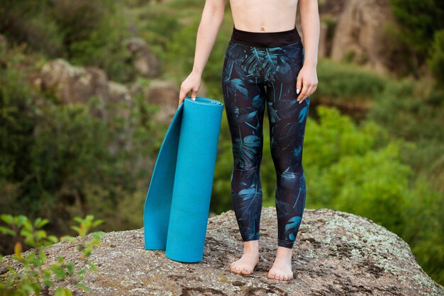 Joven deportiva con estera de yoga, de pie sobre la roca en el cañón