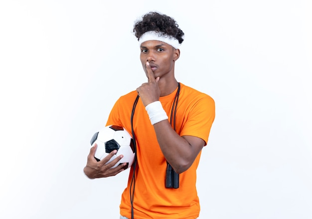 Joven deportivo afroamericano vistiendo diadema y muñequera sosteniendo la bola mostrando gesto de silencio con saltar la cuerda en el hombro