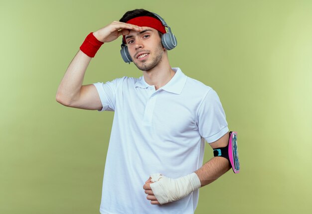 Joven deportivo en diadema con auriculares y brazalete de smartphone mirando lejos con la mano sobre la cabeza sobre verde