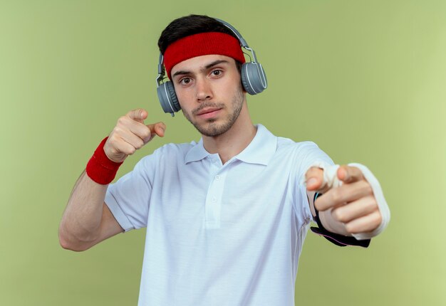 Joven deportivo en diadema con auriculares y brazalete de teléfono inteligente apuntando con el dedo a la cámara mirando confiado de pie sobre fondo verde