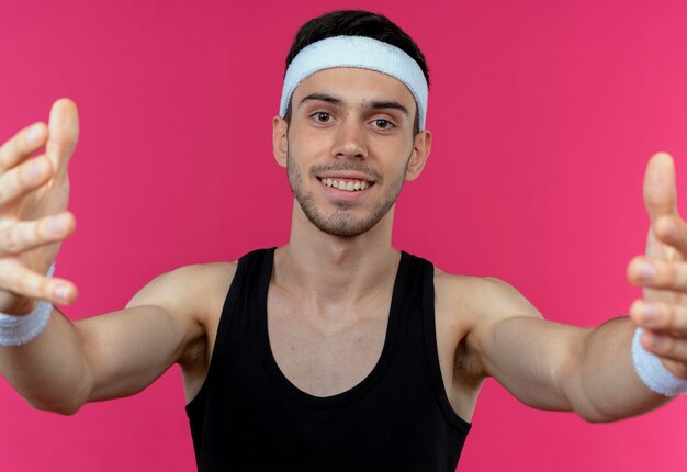 Joven deportivo en diadema haciendo gesto de bienvenida con las manos sonriendo sobre rosa