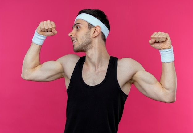 Foto gratuita joven deportivo en diadema levantando puño mostrando bíceps, concepto ganador de pie sobre fondo rosa