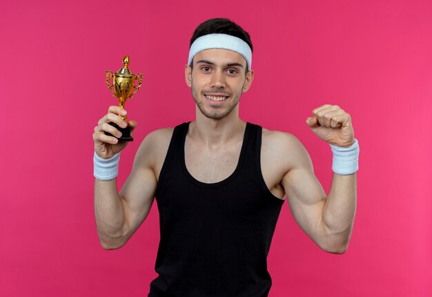 Joven deportivo en diadema con medalla de oro alrededor del cuello sosteniendo el trofeo levantando el puño y sonriendo de pie sobre la pared rosa