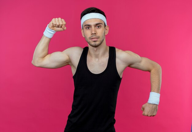 Joven deportivo en diadema posando como atleta levantando el puño mostrando bíceps sobre rosa