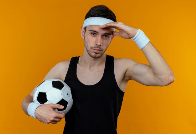 Joven deportivo en diadema sosteniendo un balón de fútbol con las manos sobre la cabeza con cara seria de pie sobre la pared naranja