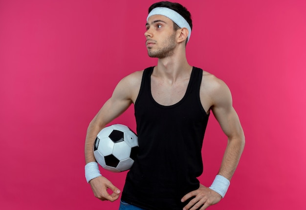 Joven deportivo en diadema sosteniendo un balón de fútbol mirando a un lado con cara seria de pie sobre la pared rosa