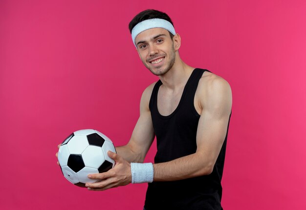 Joven deportivo en diadema sosteniendo un balón de fútbol sonriendo alegremente de pie sobre la pared rosa