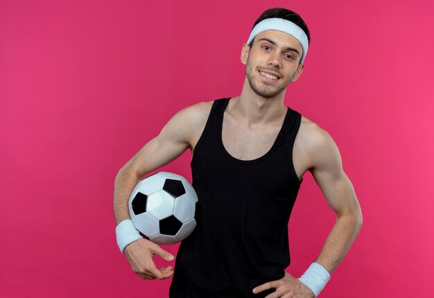 Joven deportivo en diadema sosteniendo un balón de fútbol sonriendo alegremente de pie sobre la pared rosa