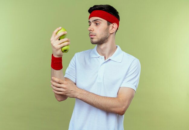 Joven deportivo en diadema sosteniendo dos manzanas verdes mirándolos con cara seria sobre verde