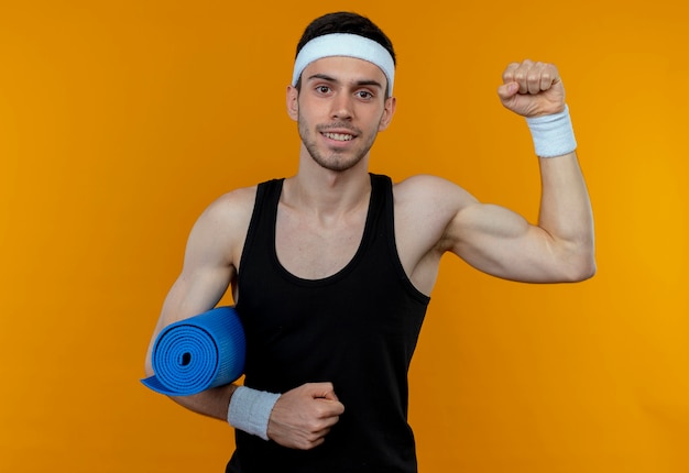 Joven deportivo en diadema sosteniendo estera de yoga levantando el puño sonriendo de pie sobre la pared naranja