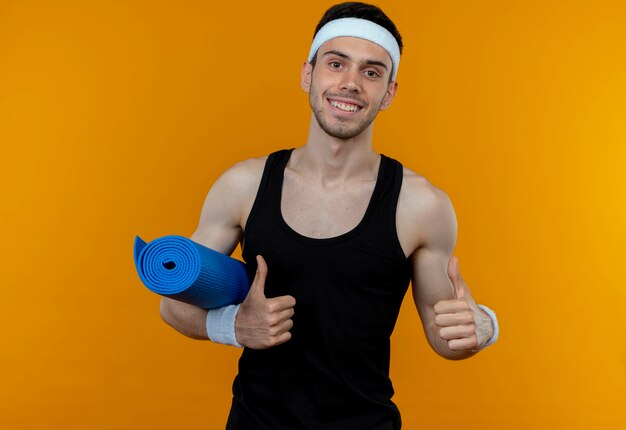 Joven deportivo en diadema sosteniendo estera de yoga sonriendo mostrando los pulgares para arriba de pie sobre la pared naranja