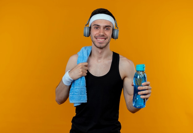 Joven deportivo en diadema con toalla en el hombro sosteniendo una botella de agua mirando a la cámara sonriendo de pie sobre fondo naranja