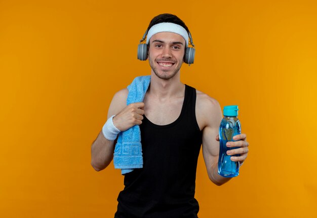 Joven deportivo en diadema con toalla en el hombro sosteniendo una botella de agua sonriendo sobre naranja