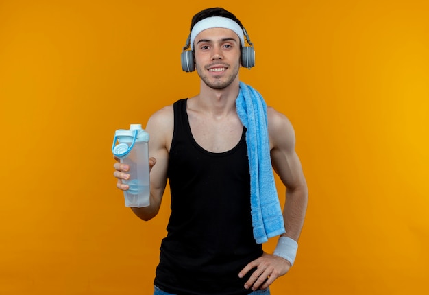 Joven deportivo en diadema con toalla en el hombro sosteniendo una botella de agua con una sonrisa en la cara sobre naranja