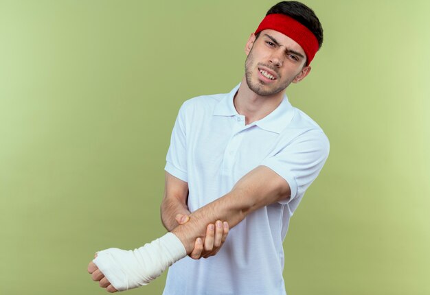 Joven deportivo en diadema tocando su mano vendada sintiendo dolor de pie sobre fondo verde