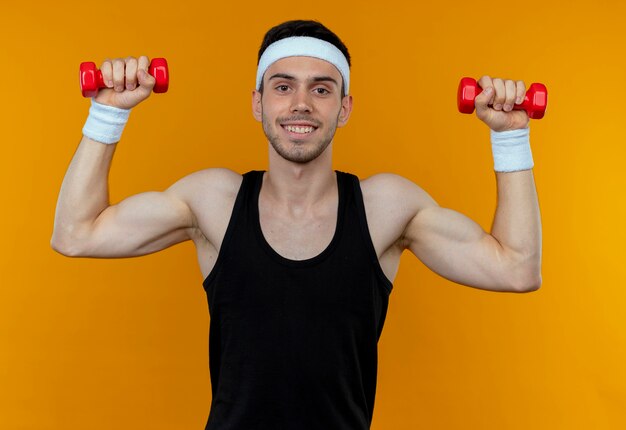 Joven deportivo en diadema trabajando con pesas sonriendo sobre naranja