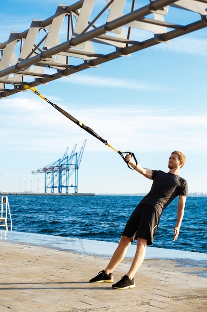 Joven deportivo entrenamiento con trx cerca del mar en la mañana.