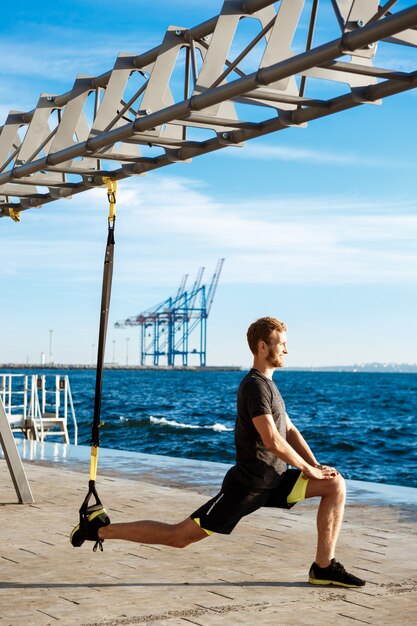 Joven deportivo entrenamiento con trx cerca del mar en la mañana.