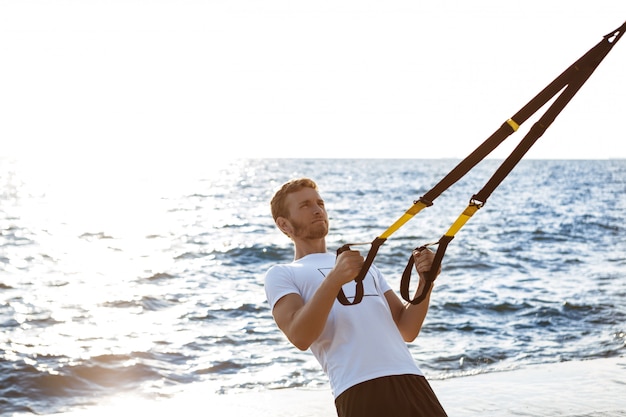 Foto gratuita joven deportivo entrenamiento con trx cerca del mar en la mañana.