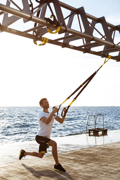 Joven deportivo entrenamiento con trx cerca del mar en la mañana.