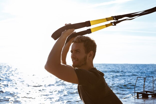 Joven deportivo entrenamiento con trx cerca del mar en la mañana.