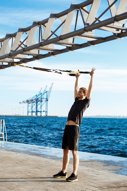 Foto gratuita joven deportivo entrenando con trx cerca del mar en la mañana