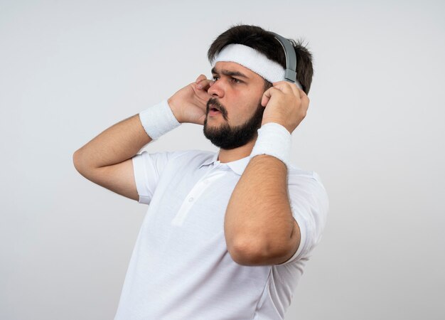 Joven deportivo mirando de lado con diadema y muñequera con auriculares aislado en la pared blanca con espacio de copia