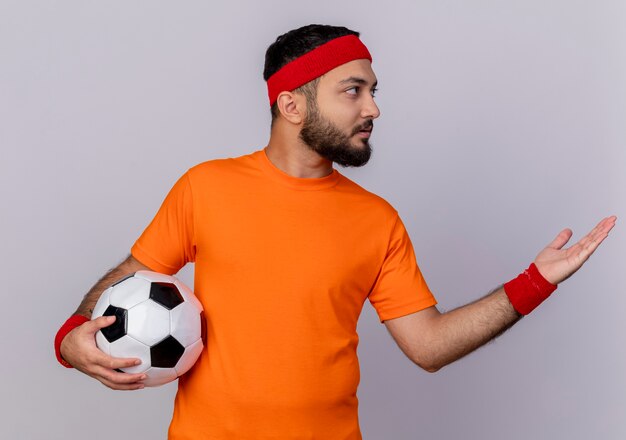 Foto gratuita joven deportivo mirando de lado con diadema y muñequera sosteniendo la bola y puntos con la mano al lado aislado sobre fondo blanco.