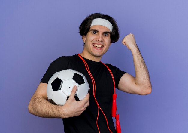 Joven deportivo con ropa deportiva y diadema con cuerda alrededor del cuello sosteniendo un balón de fútbol apretando el puño feliz y emocionado