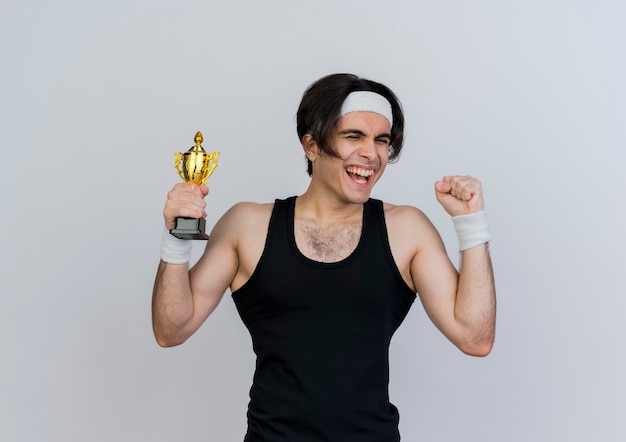 Joven deportivo con ropa deportiva y diadema sosteniendo trofeo feliz y emocionado levantando el puño feliz y emocionado de pie sobre la pared blanca