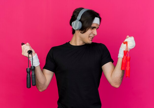 Joven deportivo vestido con ropa deportiva y diadema con auriculares sosteniendo dos cuerdas para saltar mirando confundido tratando de tomar una decisión